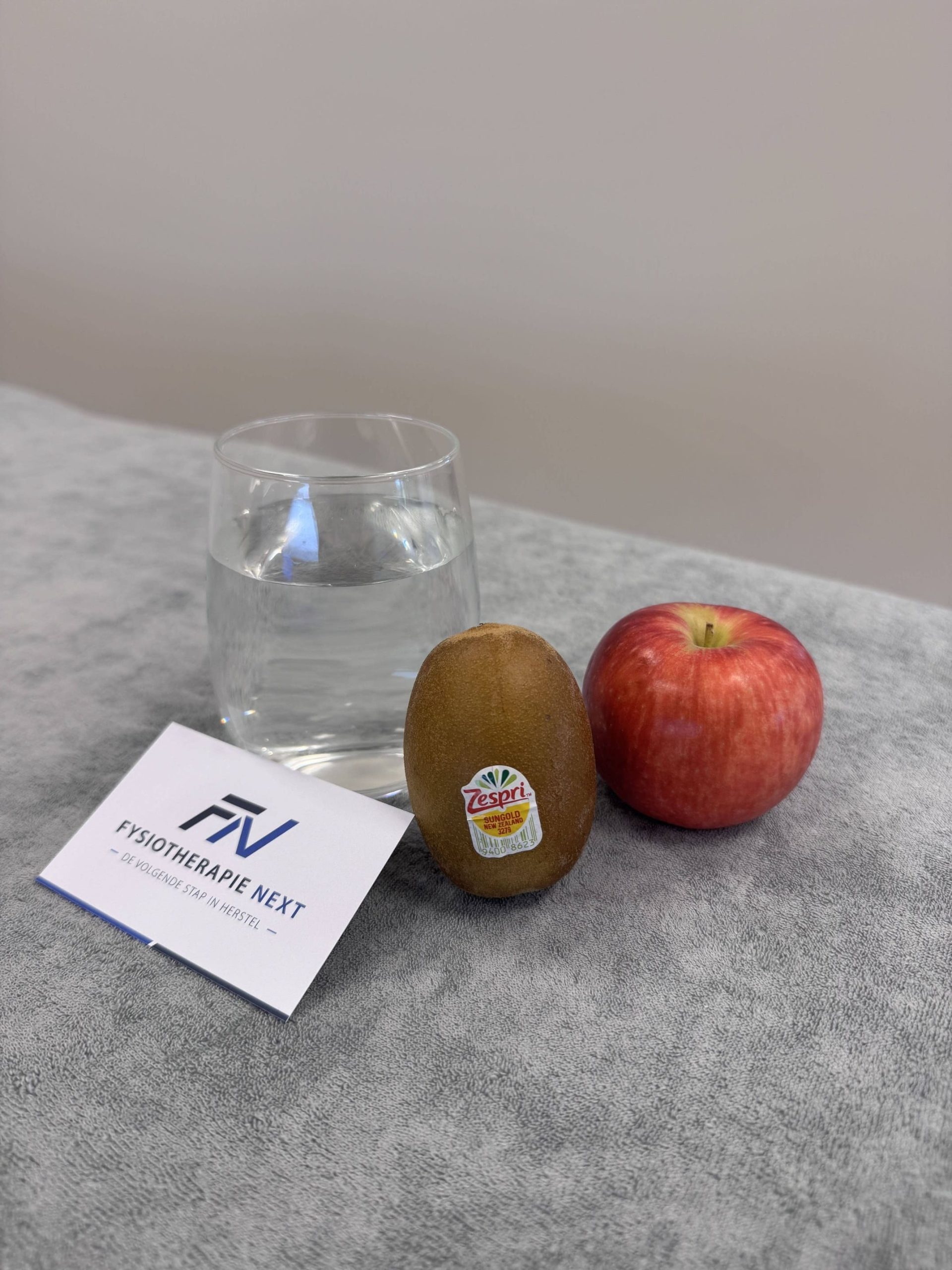 Glas water, kiwi en appel naast een kaartje van Fysiotherapie Next